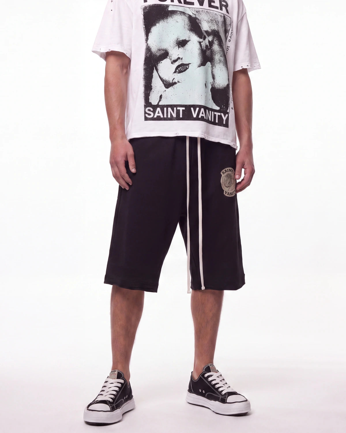 A model wearing Saint Vanty Forever brand clothing, featuring a white graphic t-shirt with a black-and-white portrait print, black drawstring shorts with white side stripes and a circular patch, and black low-top sneakers.