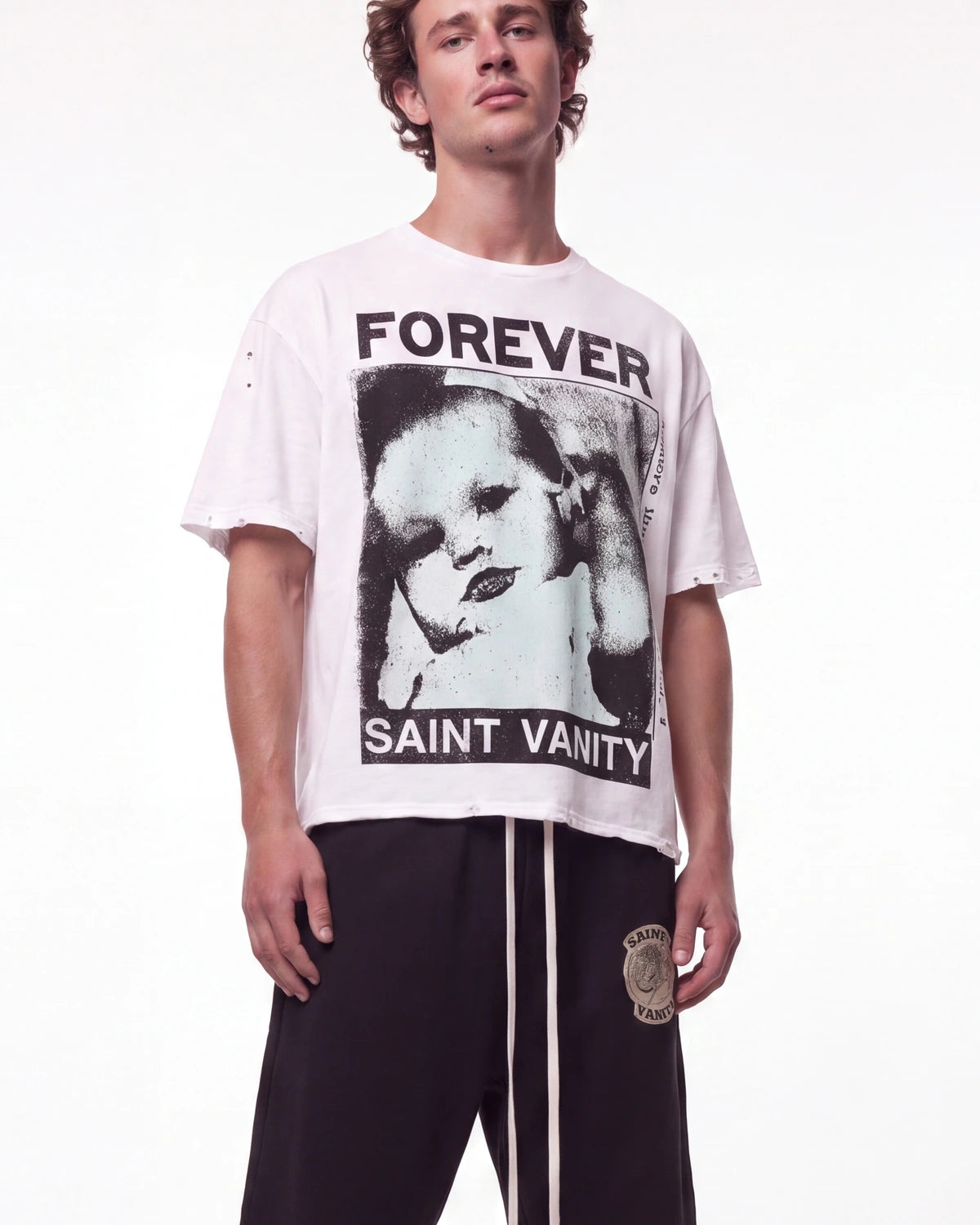 A male model wearing a white Saint Vanity graphic t-shirt with a black-and-white portrait print and distressed details, paired with black sweatpants, against a plain white studio background.