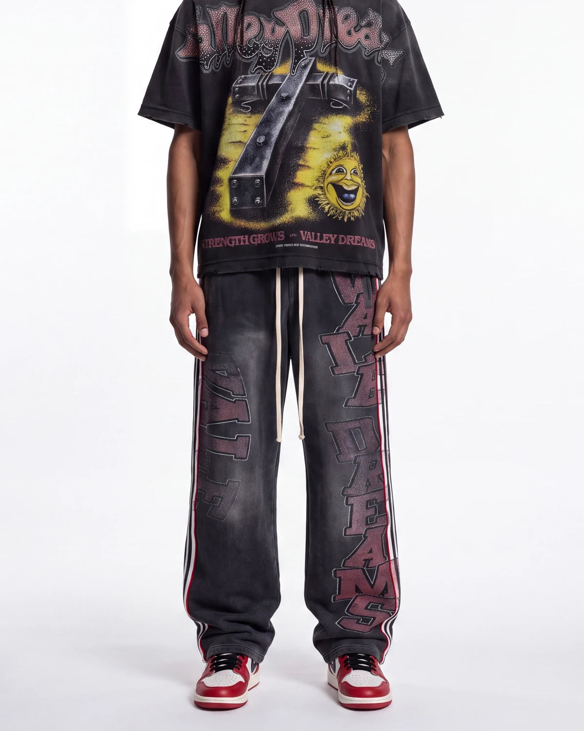 A person wearing a black graphic t-shirt and matching black distressed sweatpants with red and white side stripes and vertical lettering, paired with red and white sneakers, against a plain white studio background.