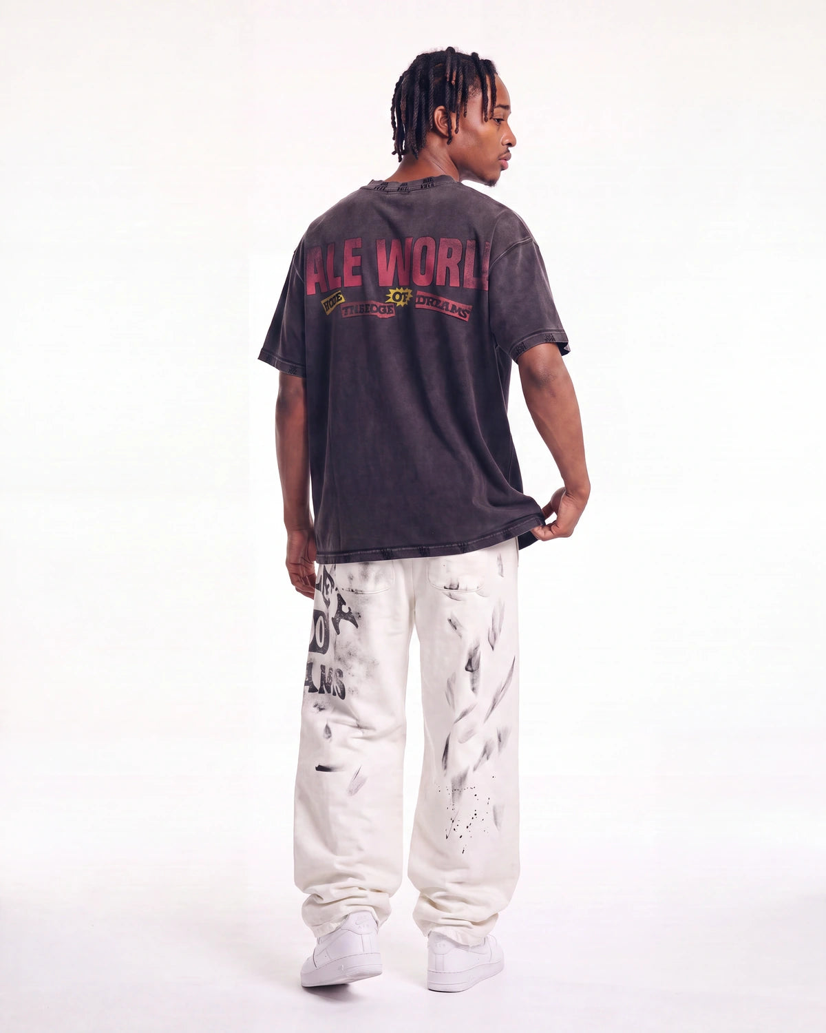 A man shown from the back wearing a faded black graphic t-shirt with 'FLE WORLD' printed in red and yellow, paired with white sweatpants and white sneakers, against a plain white studio background.