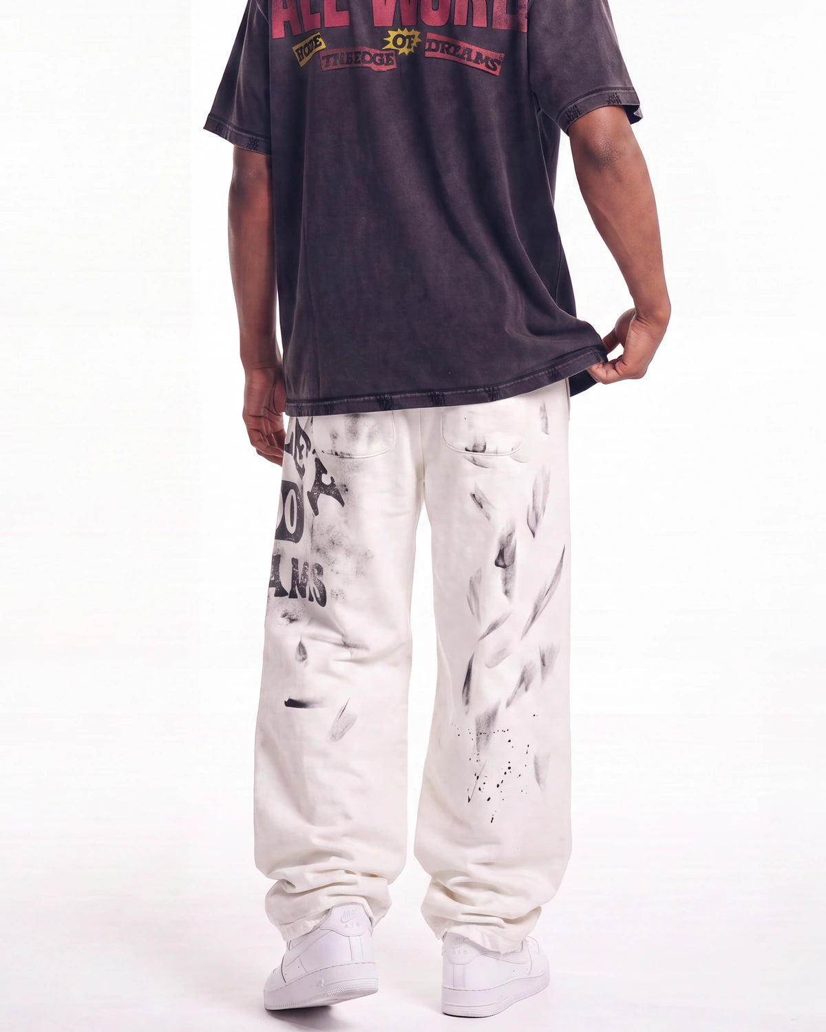 A person shown from the back wearing a faded black graphic t-shirt, off-white sweatpants with black paint splatter graphics, and white sneakers against a plain white background.
